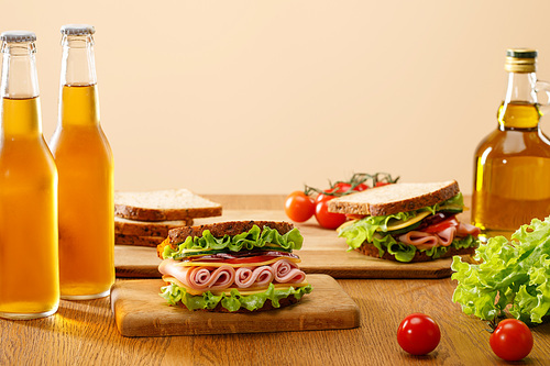 selective focus of fresh sandwiches with lettuce, ham, cheese, bacon and tomato near bottles of beer at wooden table isolated on beige