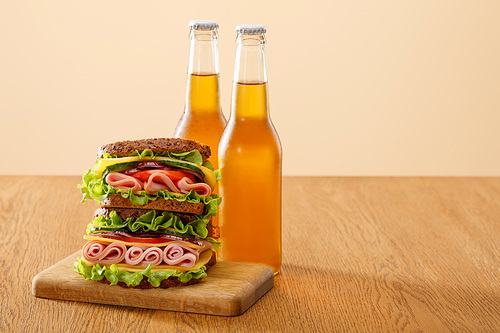 fresh sandwich with lettuce, ham, cheese, bacon and tomato near bottles of beer at wooden table isolated on beige
