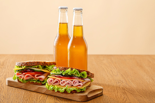 fresh sandwiches with lettuce, ham, cheese, bacon and tomato near bottles of beer at wooden table isolated on beige