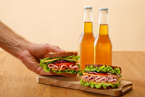 cropped view of man holding fresh sandwich with lettuce, ham, cheese, bacon and tomato near bottles of beer at wooden table isolated on beige