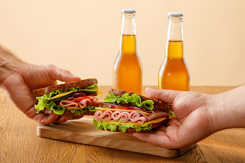 cropped view of men holding fresh sandwiches with lettuce, ham, cheese, bacon and tomato near bottles of beer at wooden table isolated on beige