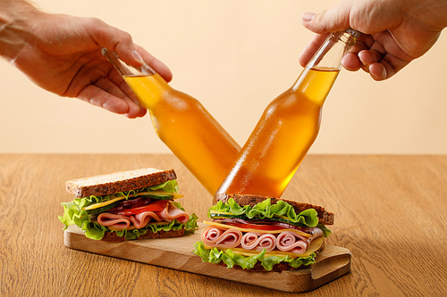 cropped view of men holding bottles of beer near fresh sandwiches with lettuce, ham, cheese, bacon and tomato near at wooden table isolated on beige