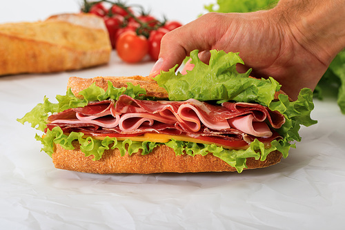 cropped view of man holding fresh sandwich with lettuce, ham, cheese, bacon and tomato on textured white background