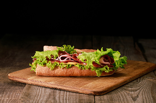 fresh sandwich with lettuce, ham, cheese, bacon and tomato on wooden cutting board isolated on black