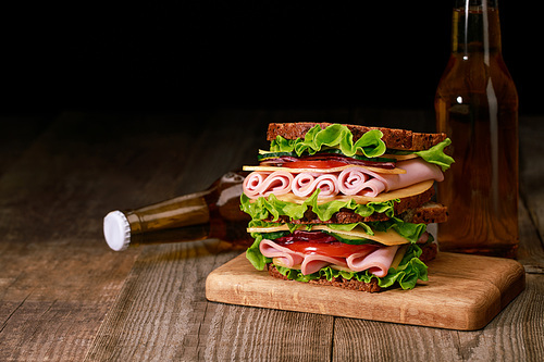 fresh sandwich with lettuce, ham, cheese, bacon and tomato on wooden cutting board near bottles of beer isolated on black