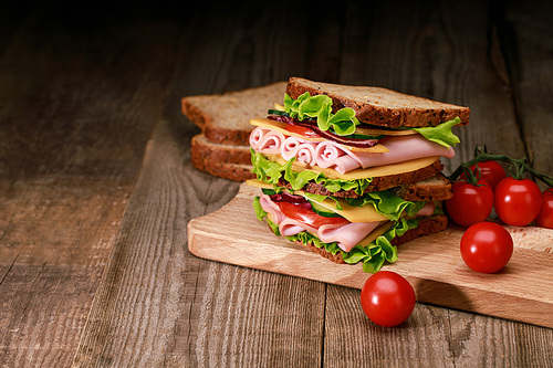 fresh sandwich with lettuce, ham, cheese, bacon and tomato on wooden cutting board with cherry tomato
