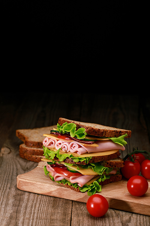 fresh sandwich with lettuce, ham, cheese, bacon and tomato on wooden cutting board with cherry tomatoes isolated on black