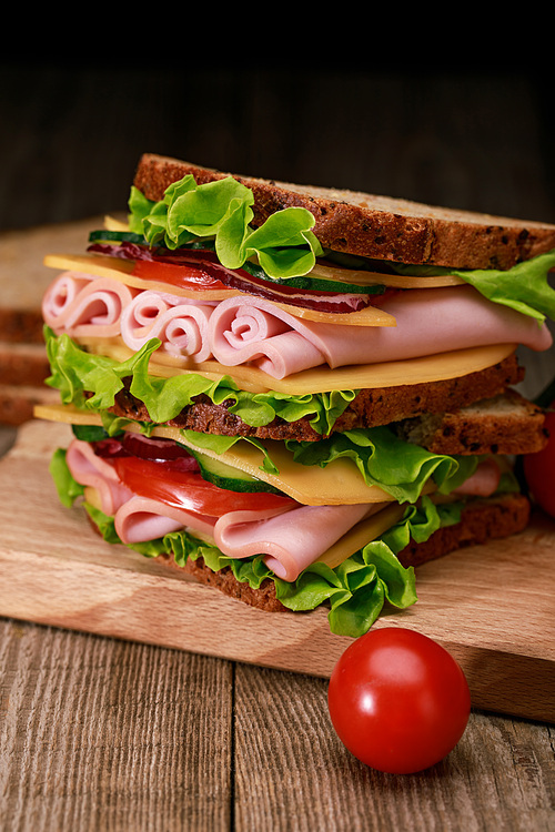 fresh sandwich with lettuce, ham, cheese, bacon and tomato on wooden cutting board with cherry tomato