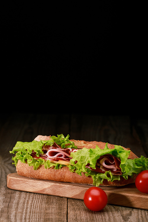 fresh sandwich with lettuce, ham, cheese, bacon on wooden cutting board with cherry tomatoes isolated on black