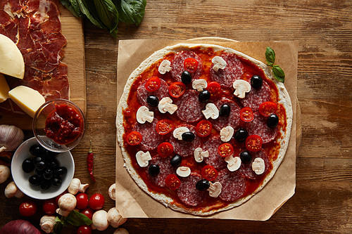 Top view of pizza with salami, mushrooms, olives, tomato sauce, parmesan, cherry tomatoes and prosciutto on wooden background