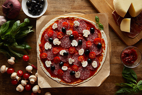 Top view of pizza with salami, mushrooms, olives, tomato sauce, vegetables, parmesan and prosciutto on wooden background