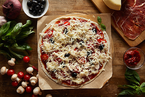 Top view of pizza on parchment paper with parmesan, vegetables, olives and salami  on wooden background
