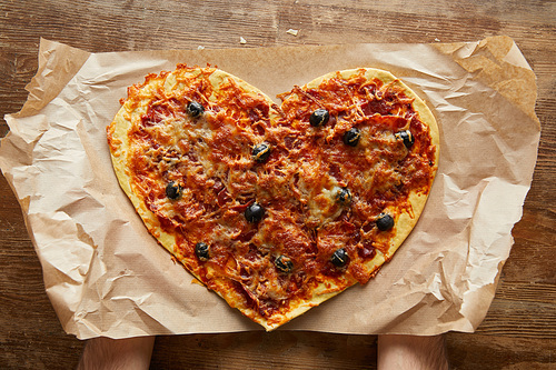 cropped view of man holding delicious italian pizza in heart shape on baking paper on wooden table