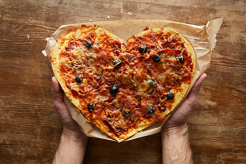 partial view of man holding delicious italian pizza in heart shape on baking paper on wooden table