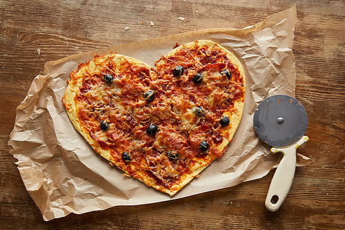 top view of delicious italian pizza in heart shape on baking paper near pizza knife on wooden table