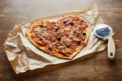 delicious italian pizza in heart shape on baking paper near pizza knife on wooden table