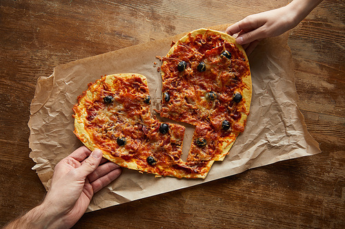 cropped view of couple holding delicious italian pizza in broken heart shape on baking paper at wooden table