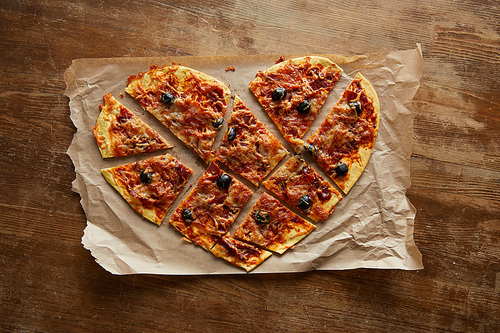 top view of delicious italian pizza in heart shape cut into pieces on baking paper on wooden table