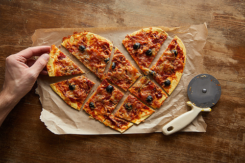 cropped view of man eating delicious italian pizza in heart shape cut into pieces on baking paper near pizza knife on wooden table