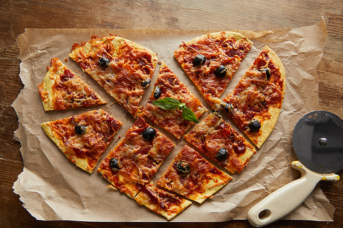 top view of delicious italian pizza in heart shape cut into pieces on baking paper near pizza knife on wooden table