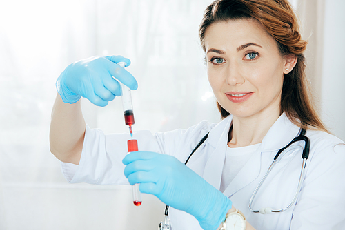 smiling doctor in latex gloves holding syringe with blood sample and test tube