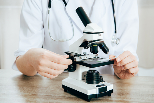 cropped view of doctor in white coat with stethoscope using microscope