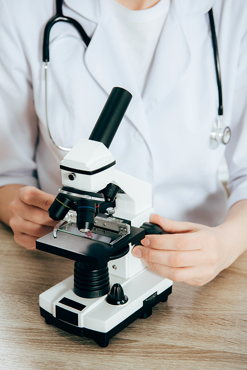 cropped view of doctor in white coat with stethoscope using microscope