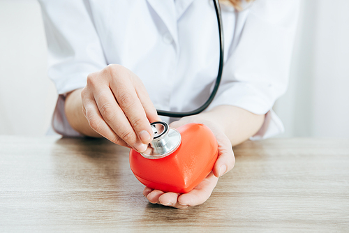 partial view of doctor with stethoscope listening plastic heart in clinic