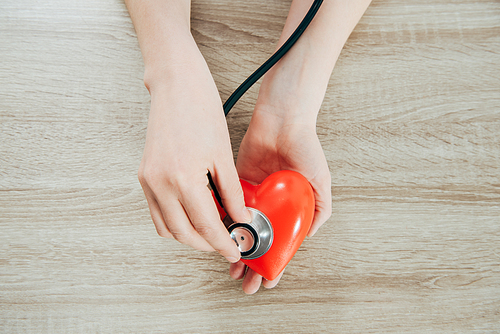 partial view of doctor with stethoscope listening plastic heart on wooden surface