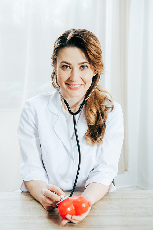 smiling doctor with stethoscope listening plastic heart in clinic