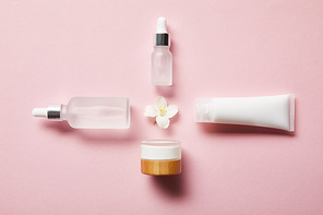 flat lay of cosmetic glass bottles, jar, cream tube with cream and jasmine flower in middle on pink