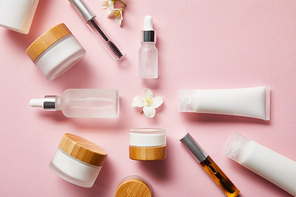 top view of jars, cream tubes with hand cream, cosmetic glass bottles, mascara bottles and jasmine flower in middle on pink