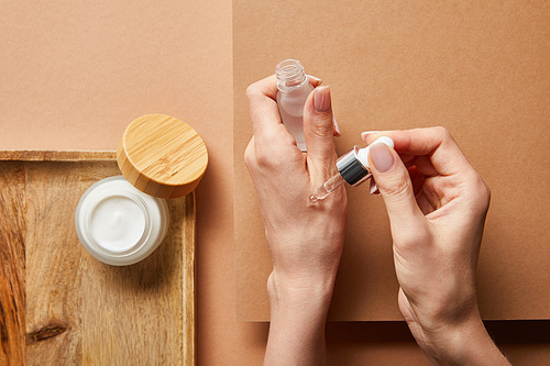 cropped view of woman holding cosmetic glass bottle with serum near open jar with cream on wooden tray on brown