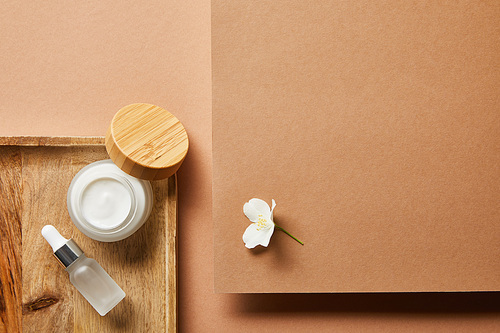 top view of open jar with cream, cosmetic glass bottle on wooden tray and jasmine flower on brown