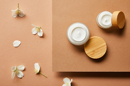 top view of open jars with cream and scattered jasmine flowers on brown