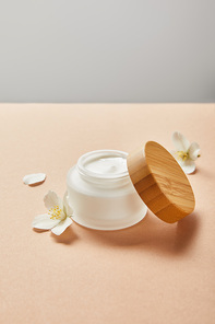 open jar with cream and wooden cap and few jasmine flowers on beige isolated on grey