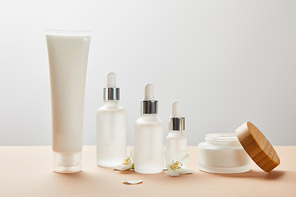 cream tube, cosmetic glass bottles, open jar with cream and few jasmine flowers on beige