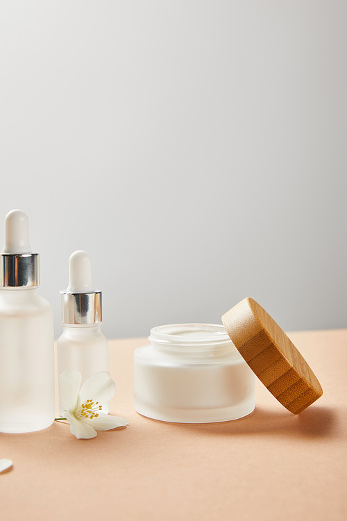 cosmetic glass bottles, open jar with cream and jasmine flower on beige