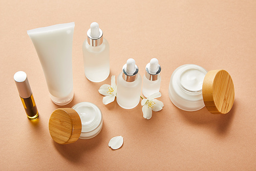 mascara bottle with natural oil, cream tube, cosmetic glass bottles, open jars with cream and jasmine flowers on beige