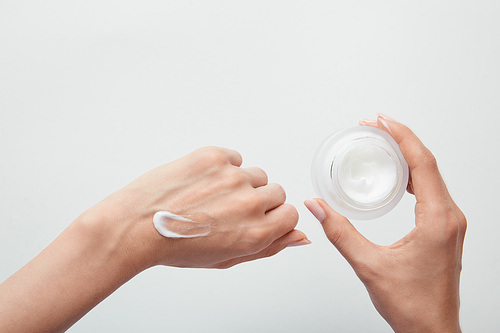 cropped view of woman applying cream on skin and holding jar isolated on white