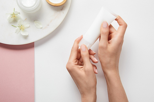 cropped view of woman holding cream tube near plate with cosmetics and flowers on white pink surface