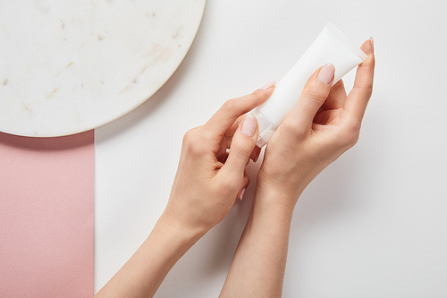 cropped view of woman holding cream tube in hands near plate on white pink surface