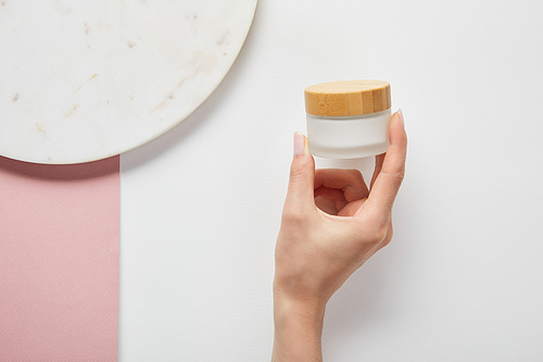 cropped view of woman holding cream in jar near plate on white pink surface