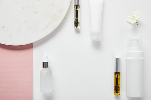 top view of mascara and glass bottles, cream tube, cosmetic dispenser near plate, jasmine on white pink surface