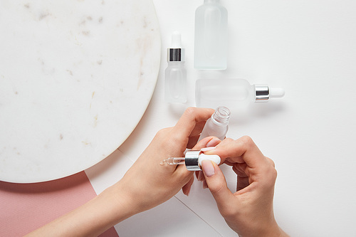 cropped view of woman applying liquid on hand near cosmetic glass bottles and plate on white pink surface