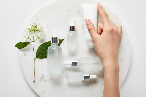 cropped view of woman hand with cream tube over plate with jasmine and cosmetic glass bottles on white surface