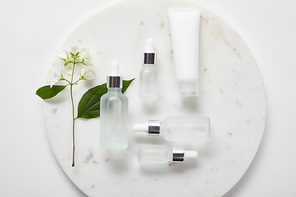 top view of cream tube and cosmetic glass bottles in plate with jasmine on white surface