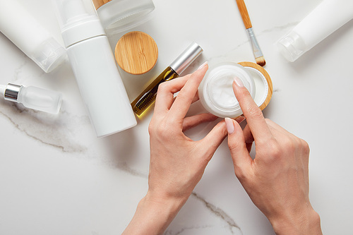 cropped view of woman holding jar with cream near tubes, cosmetic dispenser and glass bottle with eye brush on white surface