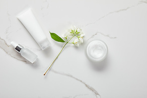 top view of cosmetic glass bottle, jar with cream, moisturizer tube and jasmine on white surface