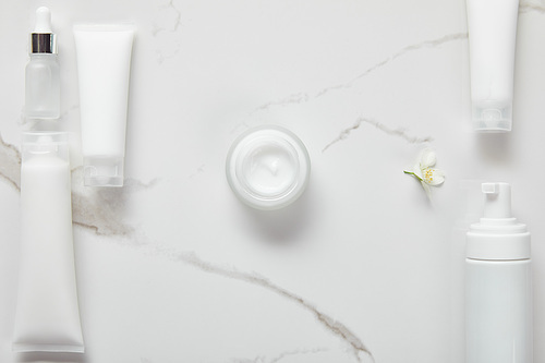 top view of cosmetic glass bottle, jar with cream, moisturizer tubes, dispenser and jasmine on white surface